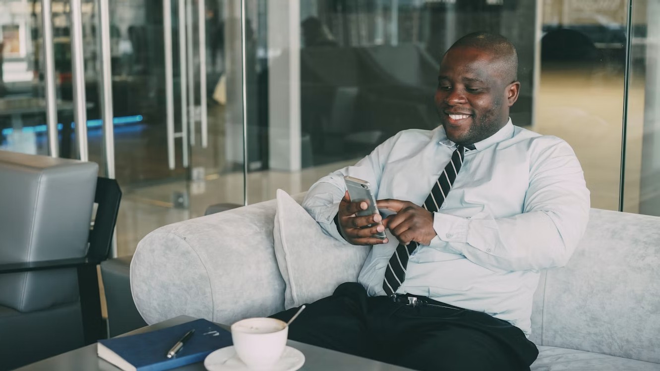 Mann in Hemd und Krawatte sitzt in einem Büro und schaut lächelnd auf sein Smartphone